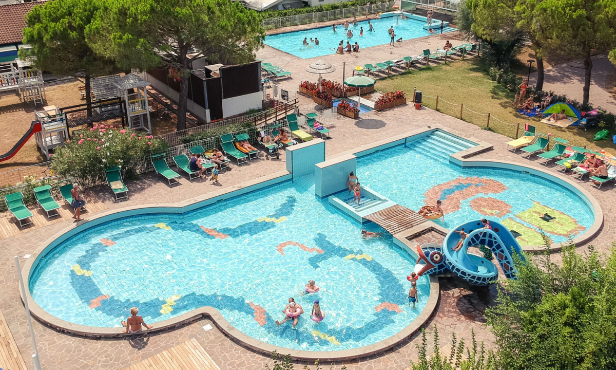 Swimming pool with slides and people lounging on sunbeds, surrounded by greenery and a playground area at Punta Spin