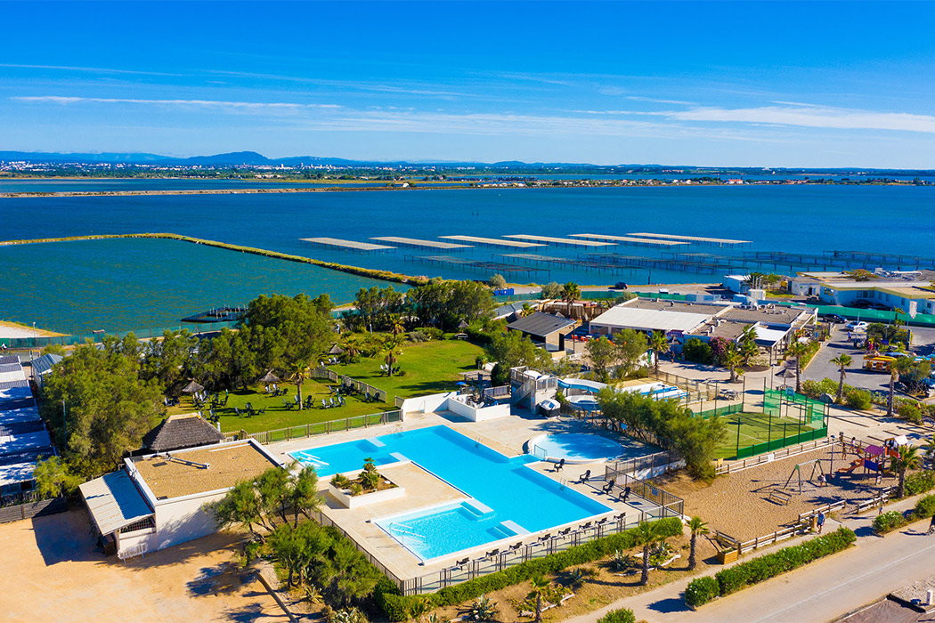 A large swimming pool and recreational area situated near a scenic body of water with surrounding greenery and buildings at Marvilla Parks Le Palavas