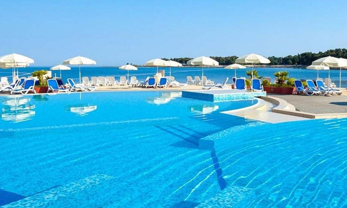 A large swimming pool beside a beach, surrounded by lounge chairs under umbrellas against a clear blue sky at Stella Plava Laguna