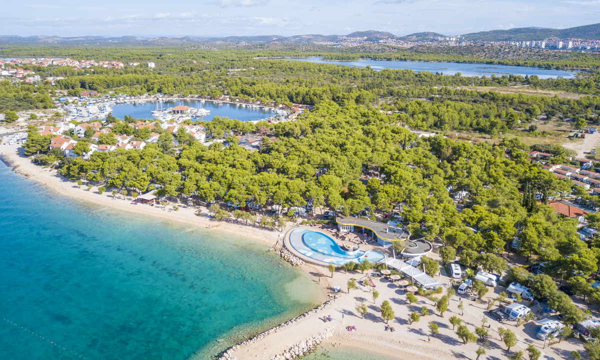 Aerial view of a beachfront camping site with dense trees, clear blue water, and scattered buildings at Amadria Park Camping Sibenik