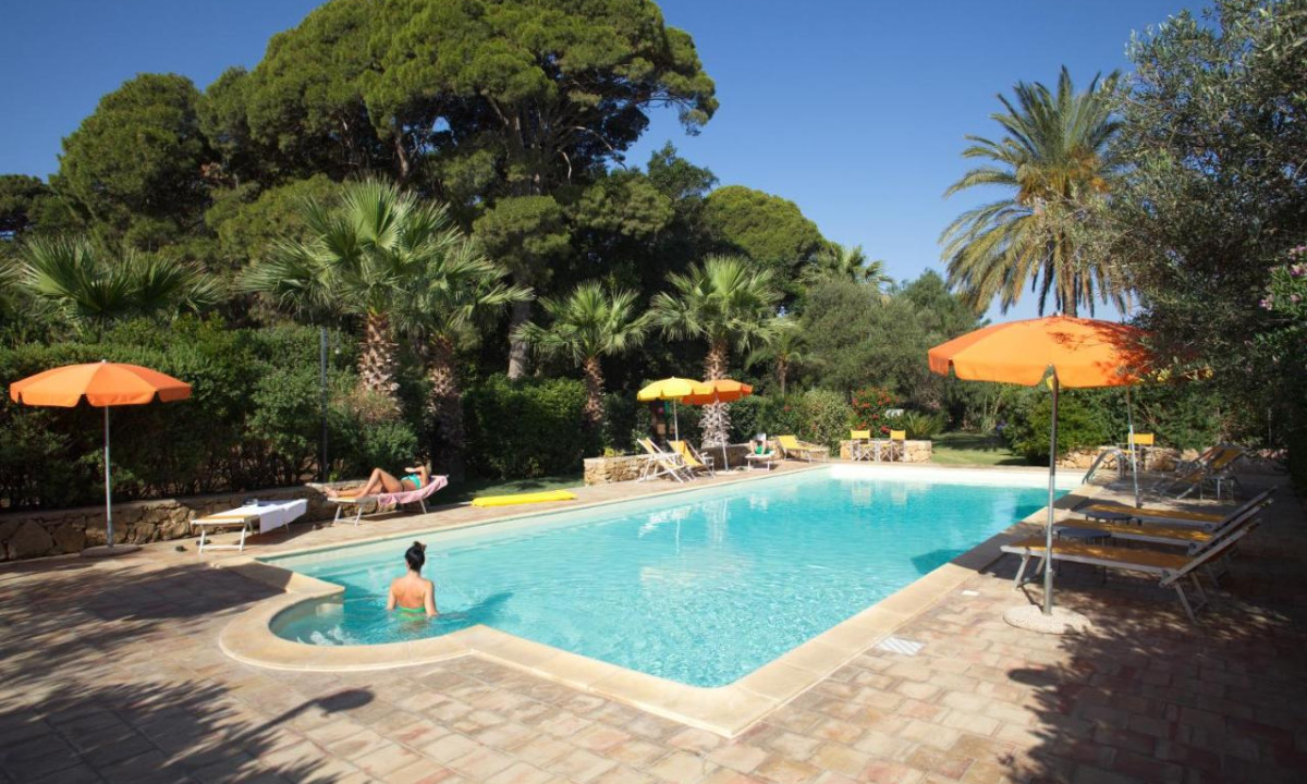 A person swims in a pool surrounded by sun loungers, orange umbrellas, and lush trees at Duca di Castelmonte