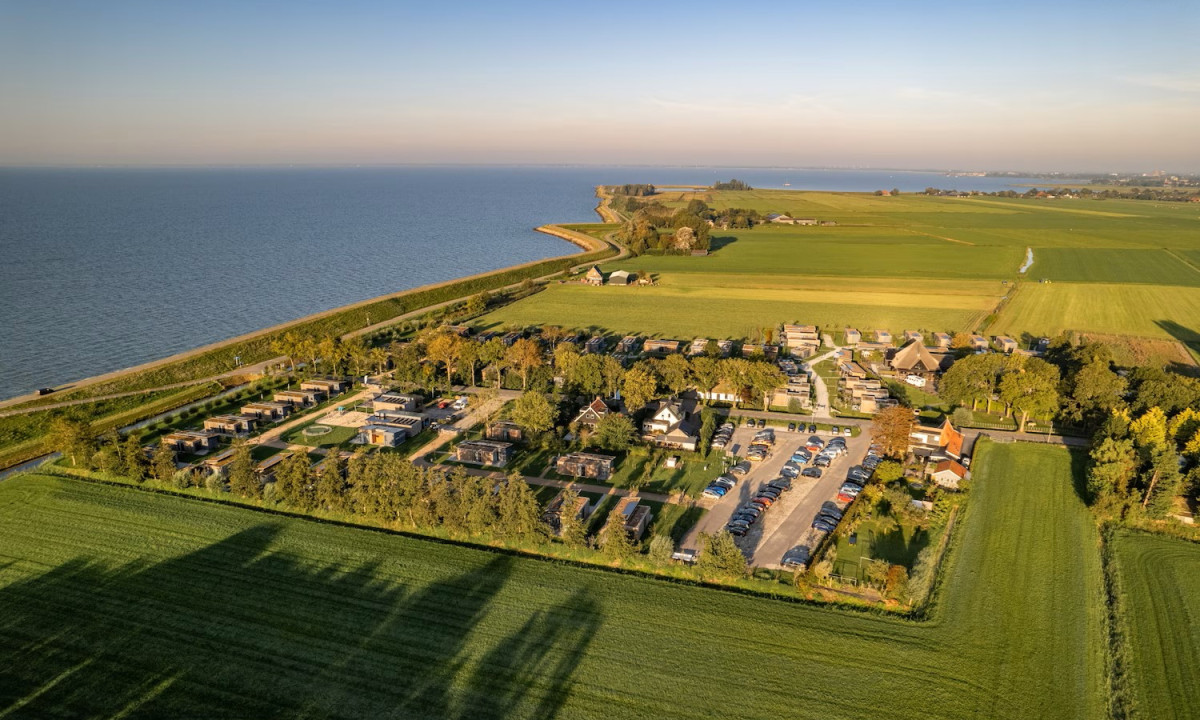 Aerial view of a coastal campground with cabins and parking, surrounded by fields and near a vast body of water at Park Wijdenes