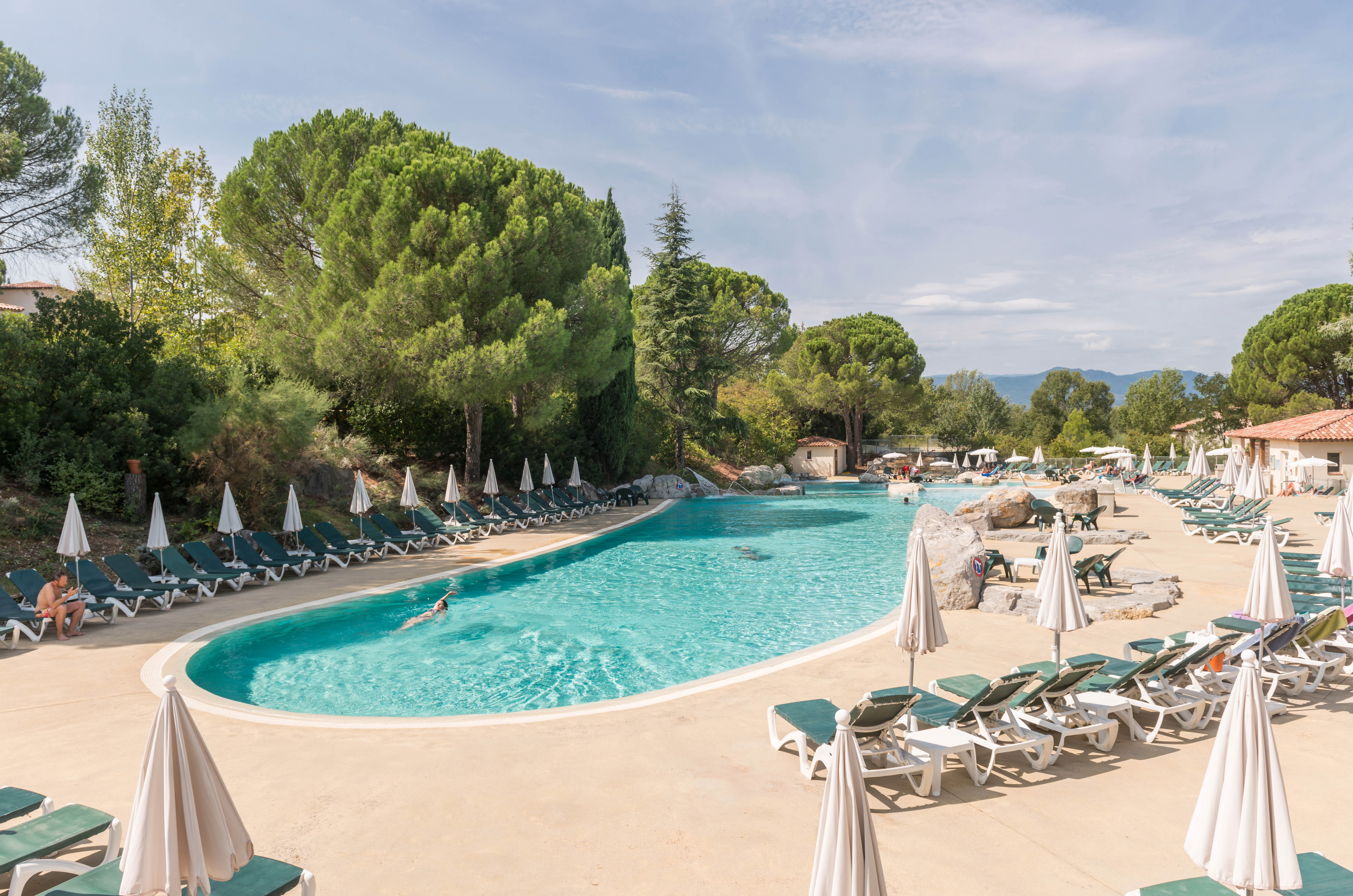 Swimming pool surrounded by lounge chairs and umbrellas, with a person swimming, in a forested campsite at Le Rouret en Ardeche