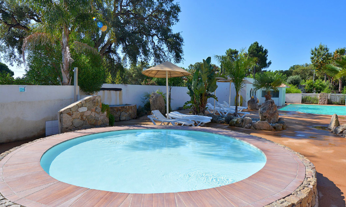 Circular wading pool remains still, surrounded by a poolside patio with loungers and umbrellas, amidst lush greenery at Via Romana