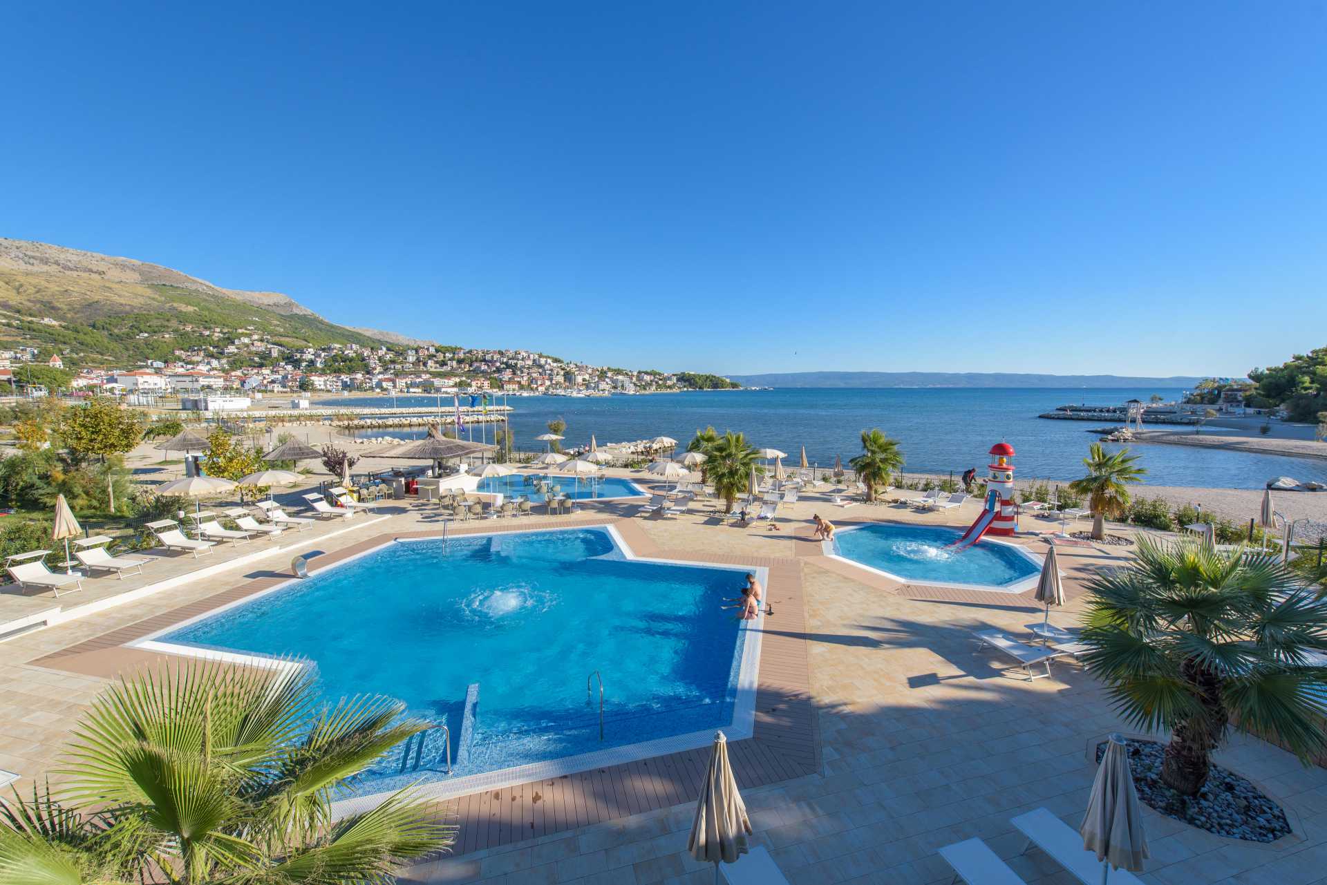 Pools with people relaxing, surrounded by palm trees, near a beach with a scenic hillside town in the background at Stobreč Split