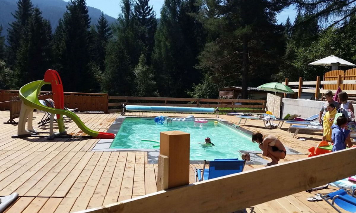 Children swimming in a small pool surrounded by people on a wooden deck with mountain scenery at Fiemme Village