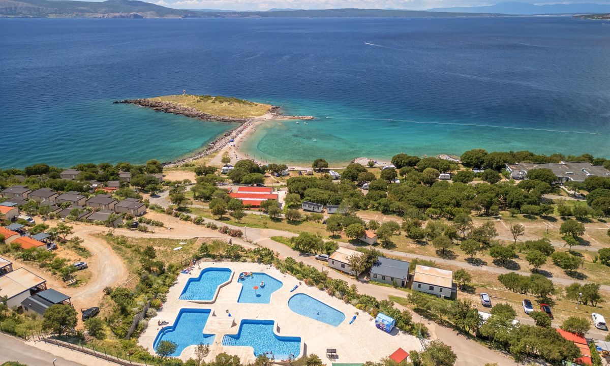Swimming pools filled with people, surrounded by trees and cabins, overlook a rocky beach and expansive ocean at Klenovica