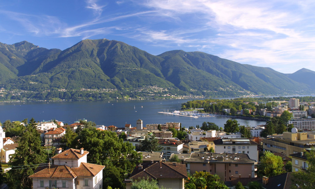 Buildings overlook a serene lake with surrounding lush green mountains under a partly cloudy blue sky at Lido di Monvalle