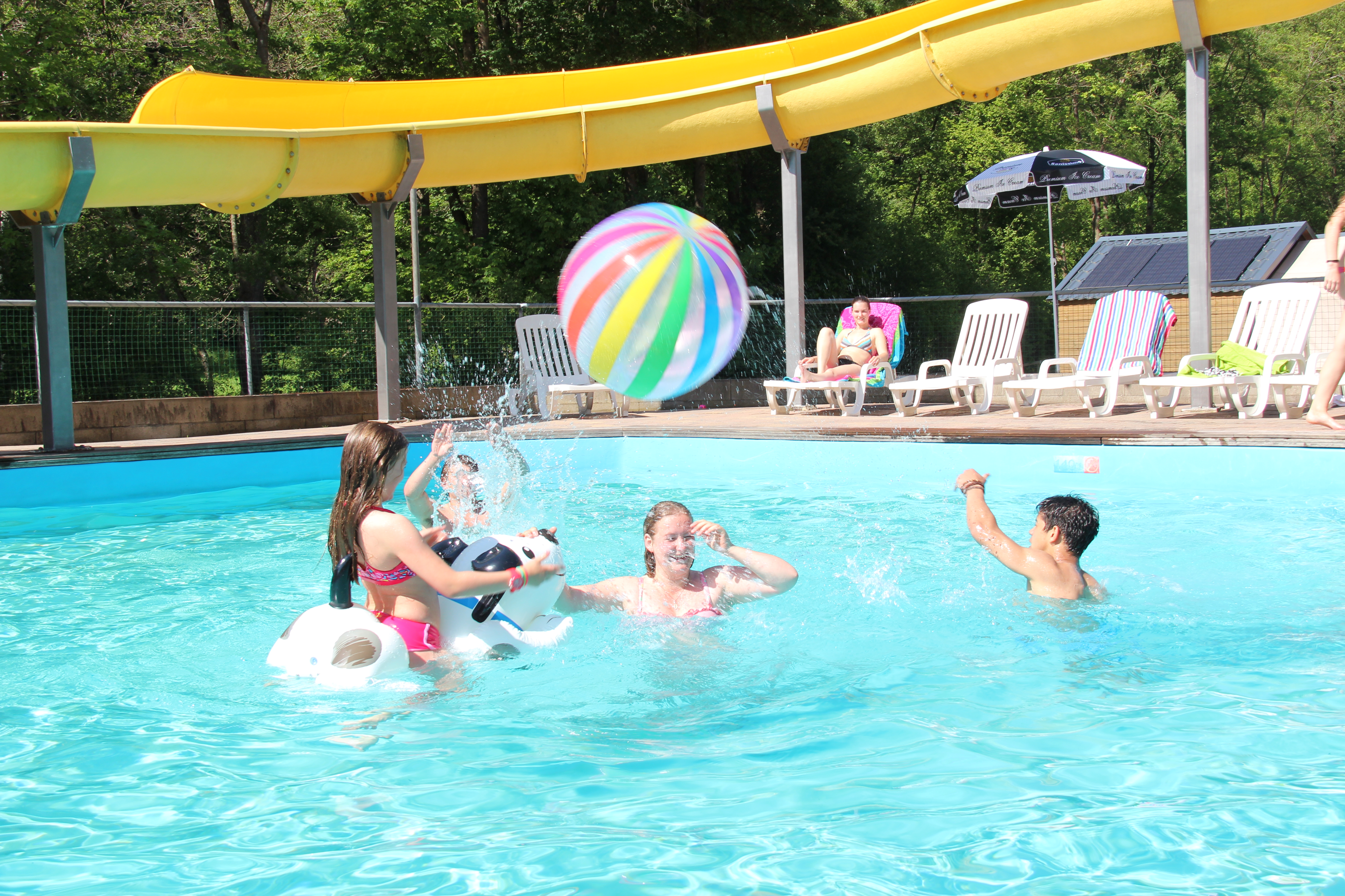 People playing with a beach ball in a swimming pool, with lounge chairs and a yellow water slide nearby at Polleur