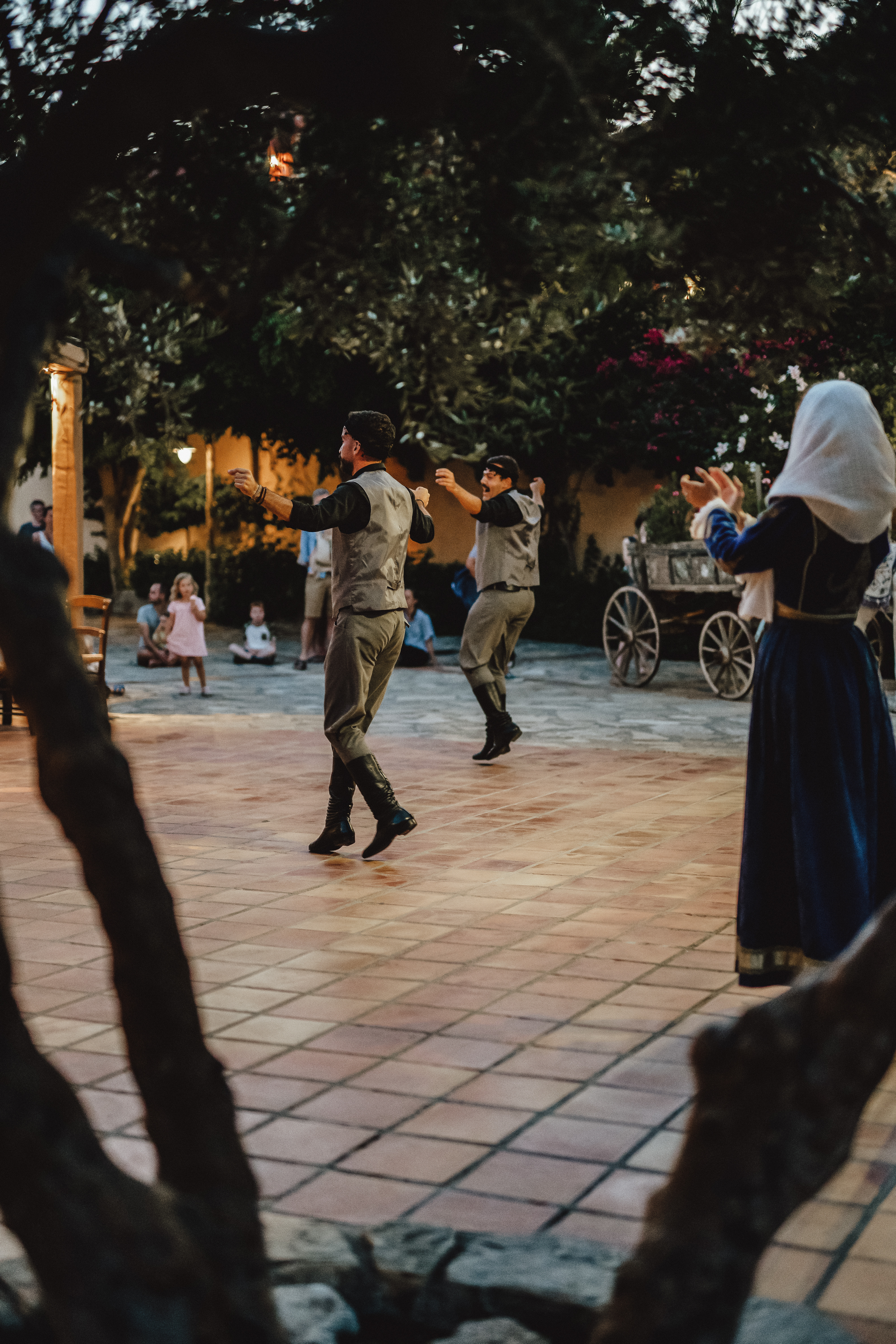 Dancers perform in traditional attire, watched by an audience on a tiled courtyard with trees and a wooden cart at Candia Park Village