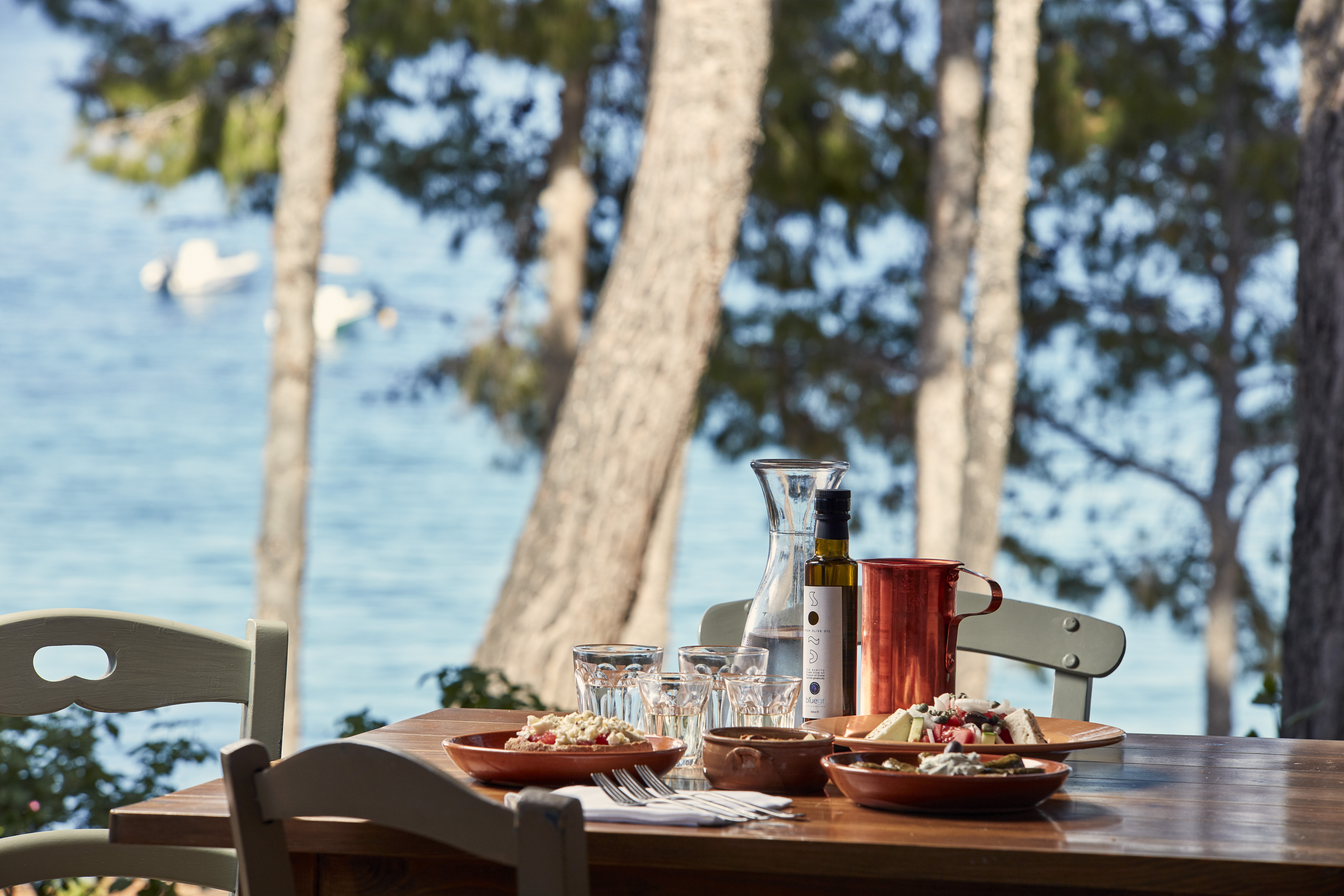 Plates of food and drinks on a wooden table surrounded by trees with a view of the sea at Candia Park Village