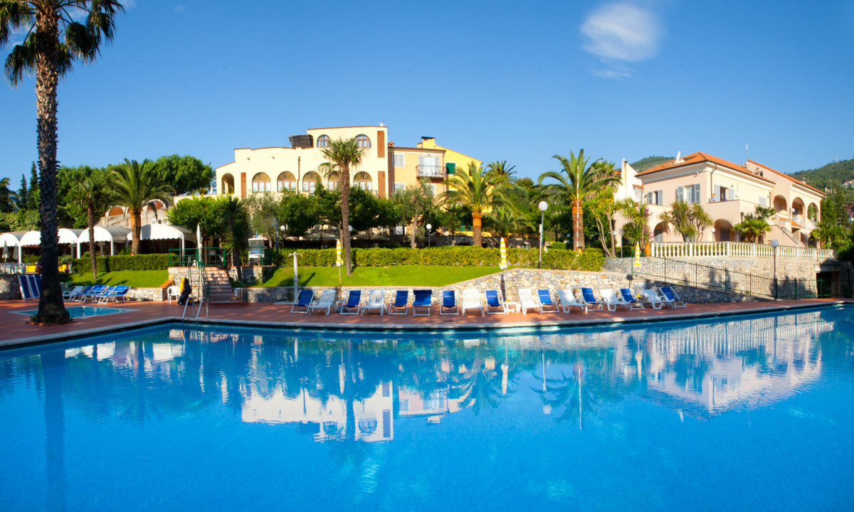 Large swimming pool reflecting the surrounding resort buildings and lush greenery in a clear, sunny day at Pian Dei Boschi