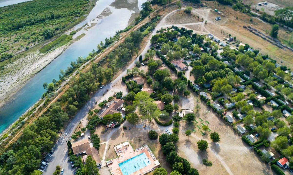 Aerial view of a swimming pool surrounded by buildings and trees, adjacent to a river and campsite at Marvilla Parks Les Rives du Luberon