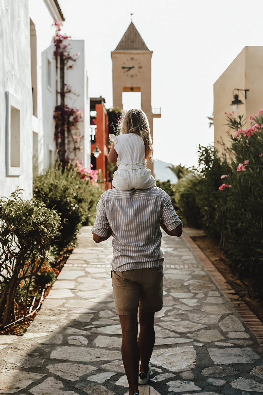 Man carrying child on shoulders, walking along a stone path between buildings with clock tower in background at Candia Park Village