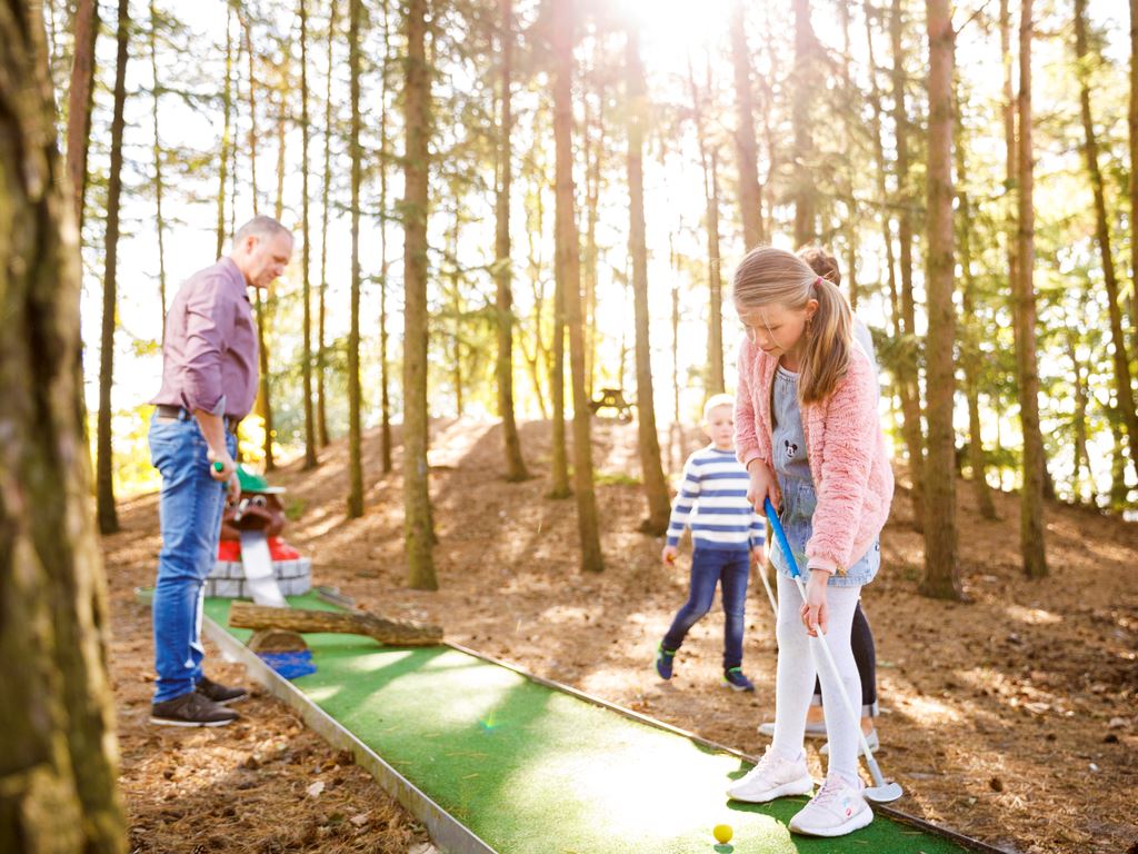 Child putting a golf ball at a mini-golf course, surrounded by trees and family, with sunlight filtering through the forest at Landal Dwergter Sand