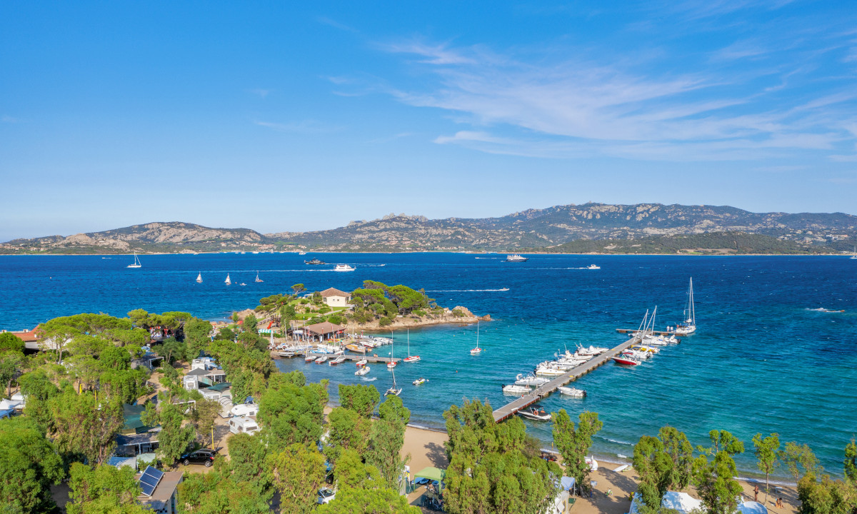 Boats are docked along a pier in clear blue water, surrounded by lush trees and distant mountains at Isuledda