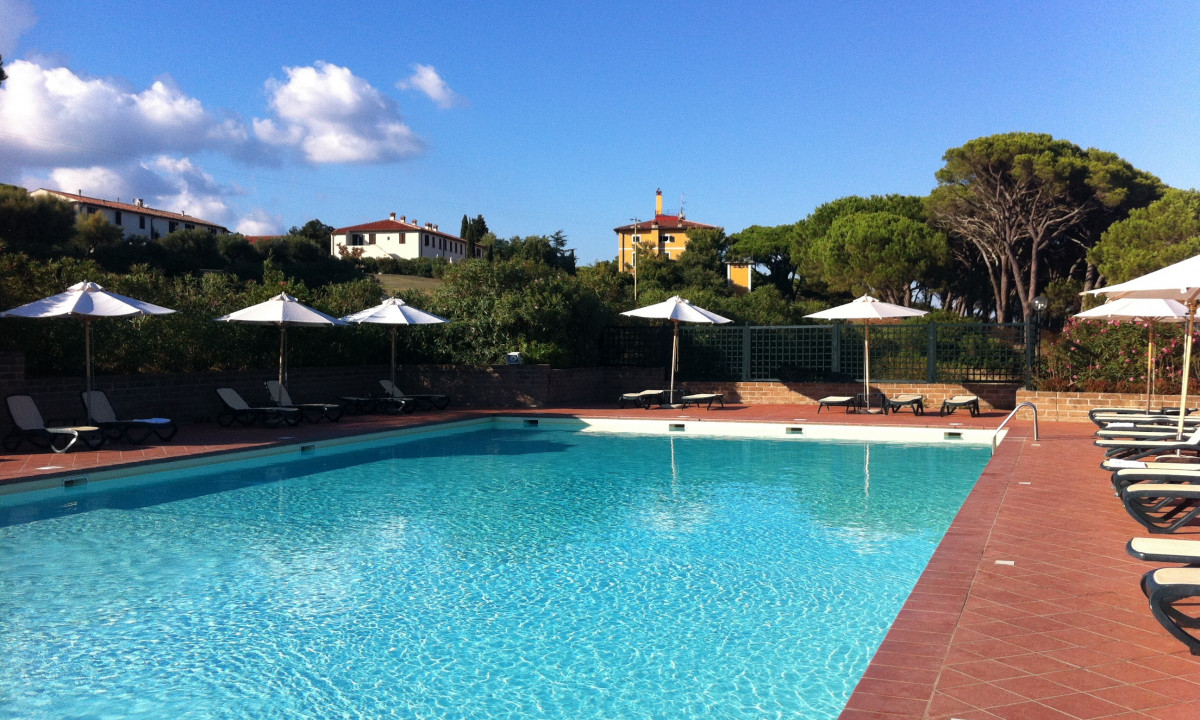 A swimming pool with lounge chairs and parasols, surrounded by greenery and buildings, under a clear blue sky at Tenuta delle Ripalte