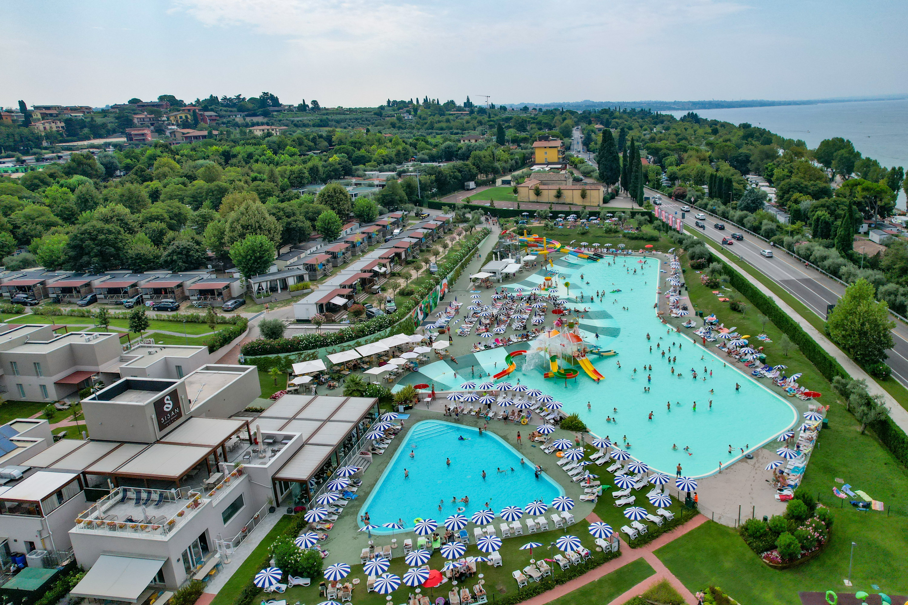 Pools with water slides are surrounded by people lounging under umbrellas, near modern buildings and lush greenery at Cisano/San Vito