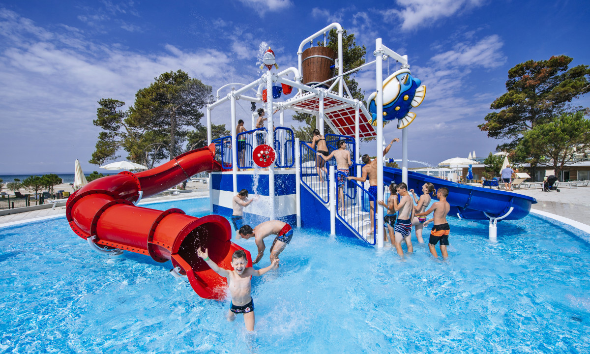 Children play in a colorful aquatic playground with slides and water features, surrounded by trees and beach at Zaton Holiday Resort