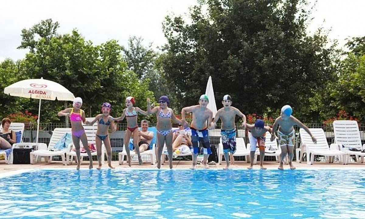 Children jump into a swimming pool, holding hands, in a vibrant outdoor setting with sunbathing adults and green trees at Okay Lido