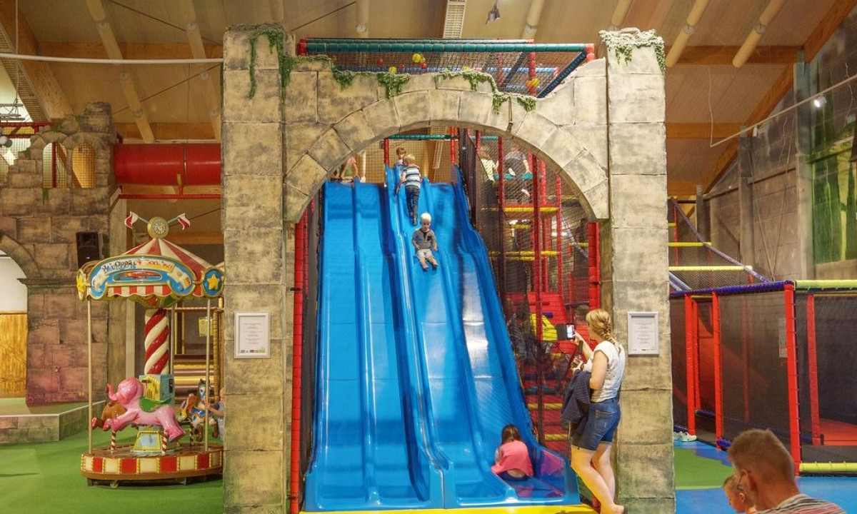 Children sliding on blue slides in an indoor playground with a stone archway and a carousel at Landal Sonnenberg