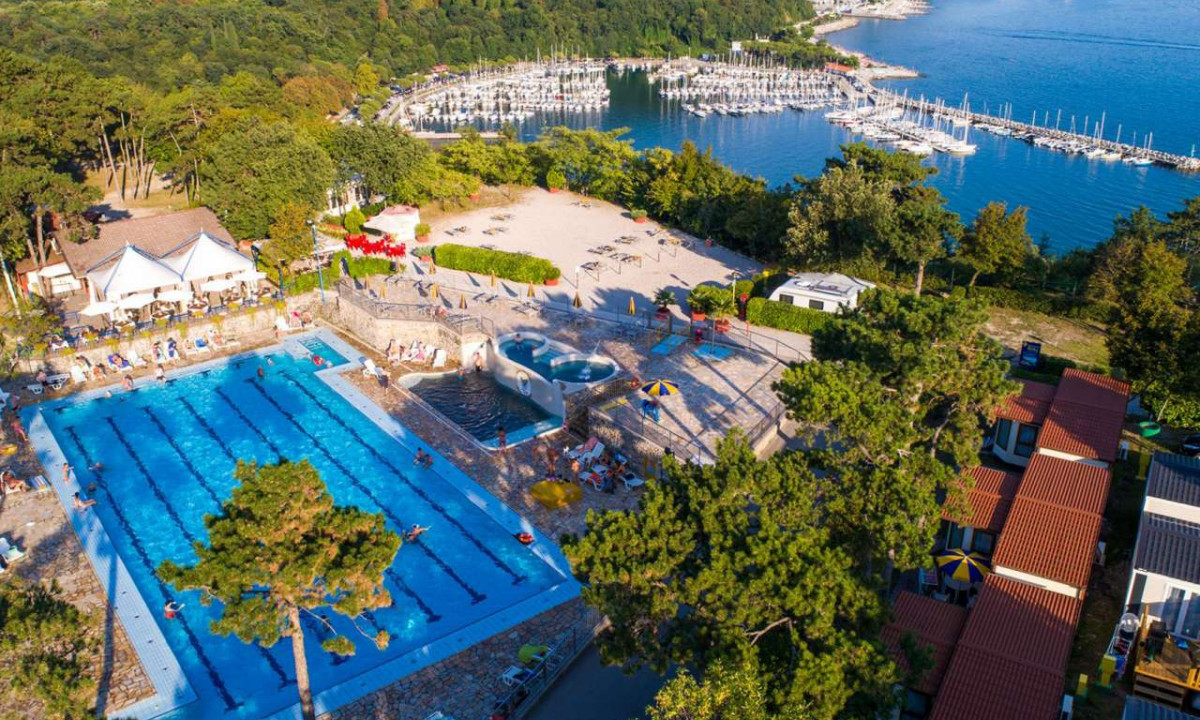 Pool packed with swimmers, flanked by lounging deck chairs and surrounded by lush trees and a lakeside marina at Camping Mare Pineta