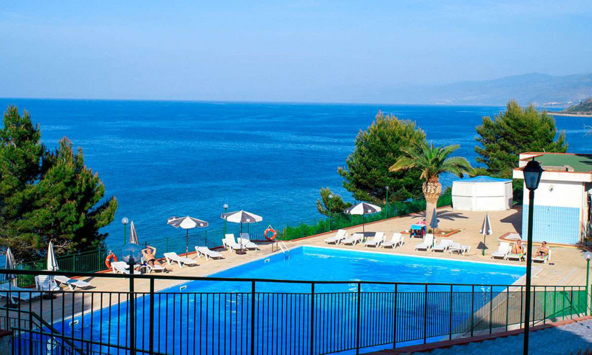 Poolside loungers sit under umbrellas by a clear blue pool, overlooking a vast ocean with distant cliffs and lush greenery at Rais Gerbi