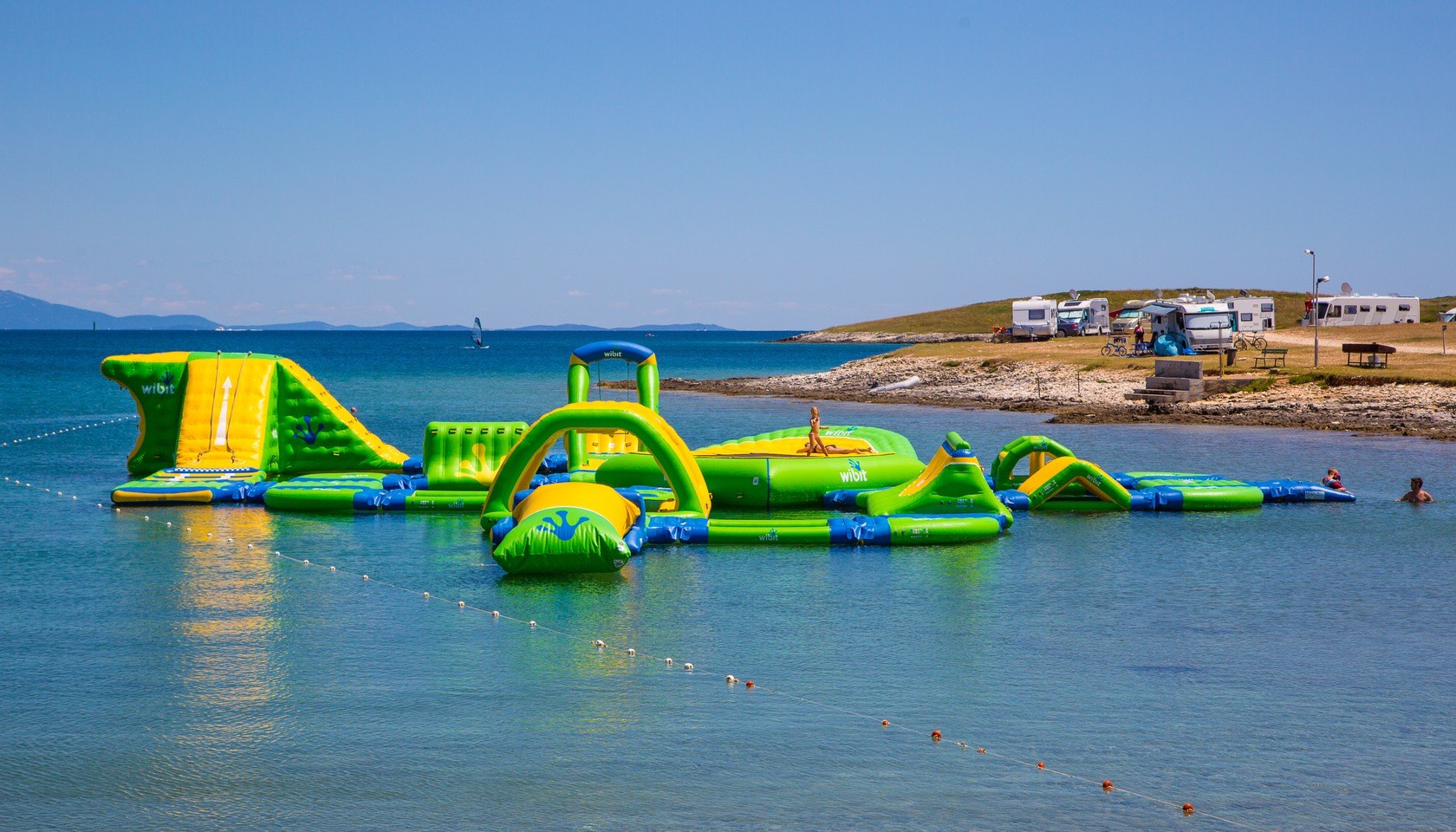 Inflatable water park floating on a calm sea with campers and RVs on the distant shore at Stupice
