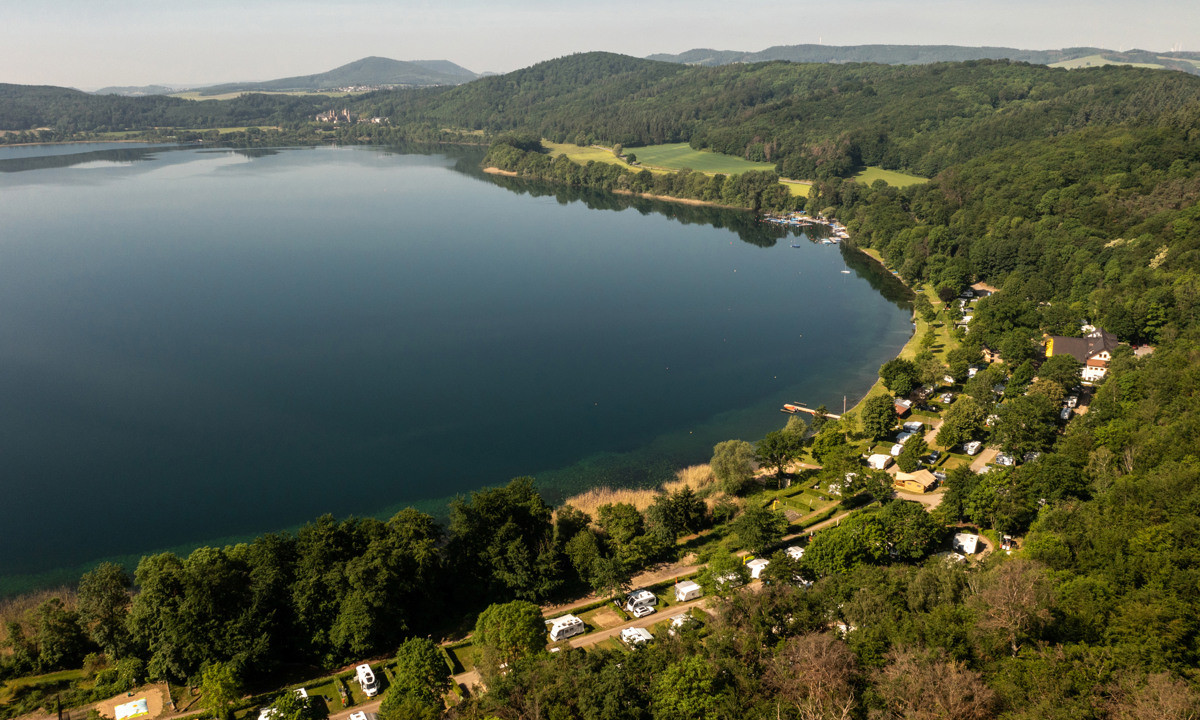 Lake reflecting landscape with trees and hills, surrounded by campsite with tents and caravans at Laacher See