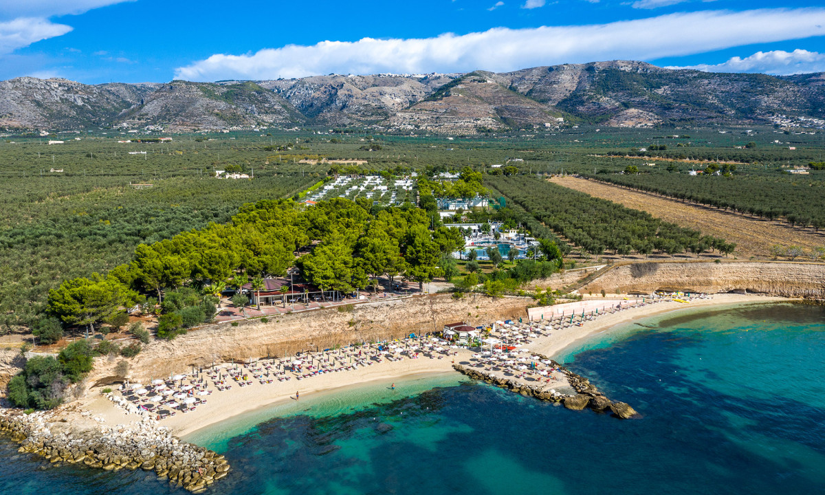 Beachgoers sunbathe on sandy beach by aquamarine waters, surrounded by green trees and mountainous farmland at Village Baia del Monaco