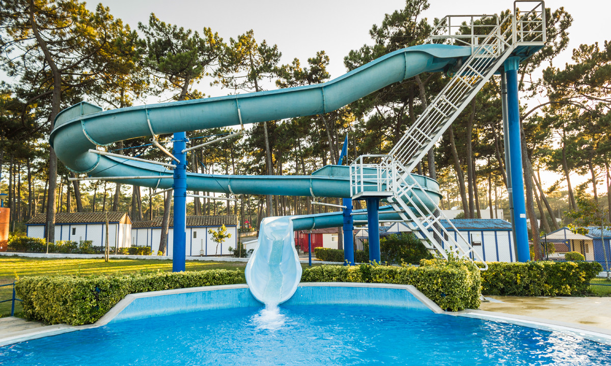 A large water slide curves down into a clear blue pool, surrounded by trees and small buildings at Sao Pedro de Moel