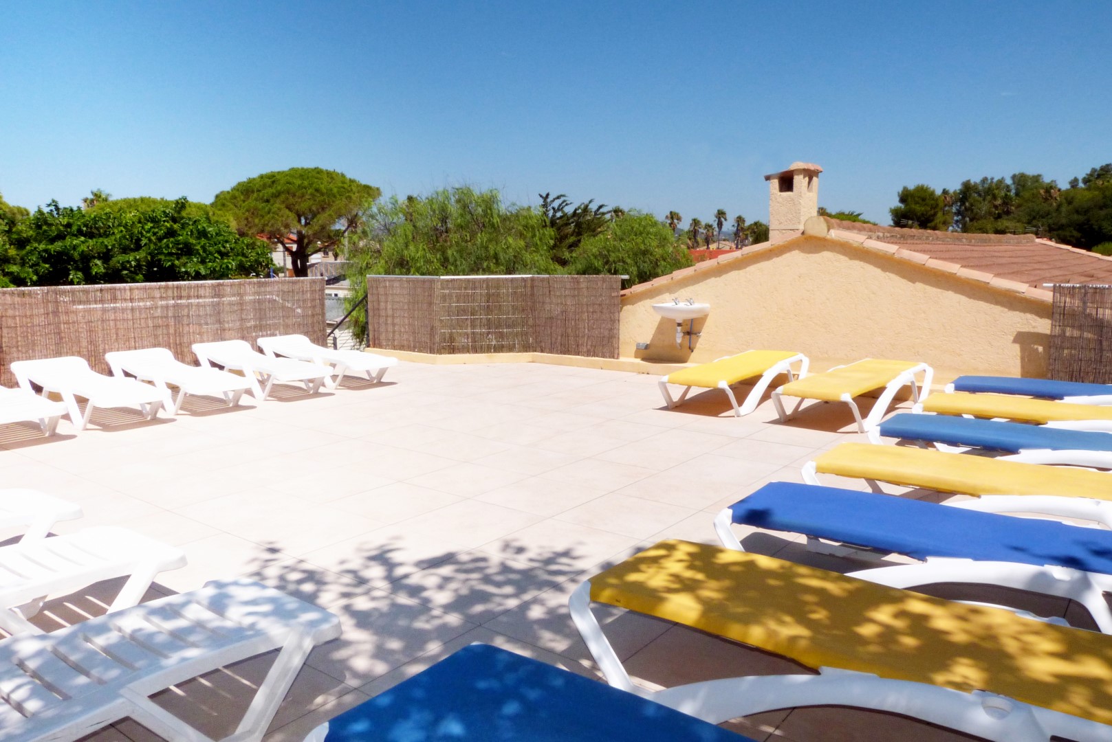 Sun loungers, aligned under blue skies, on a sunny rooftop terrace with trees and buildings in the background at La Bergerie Plage