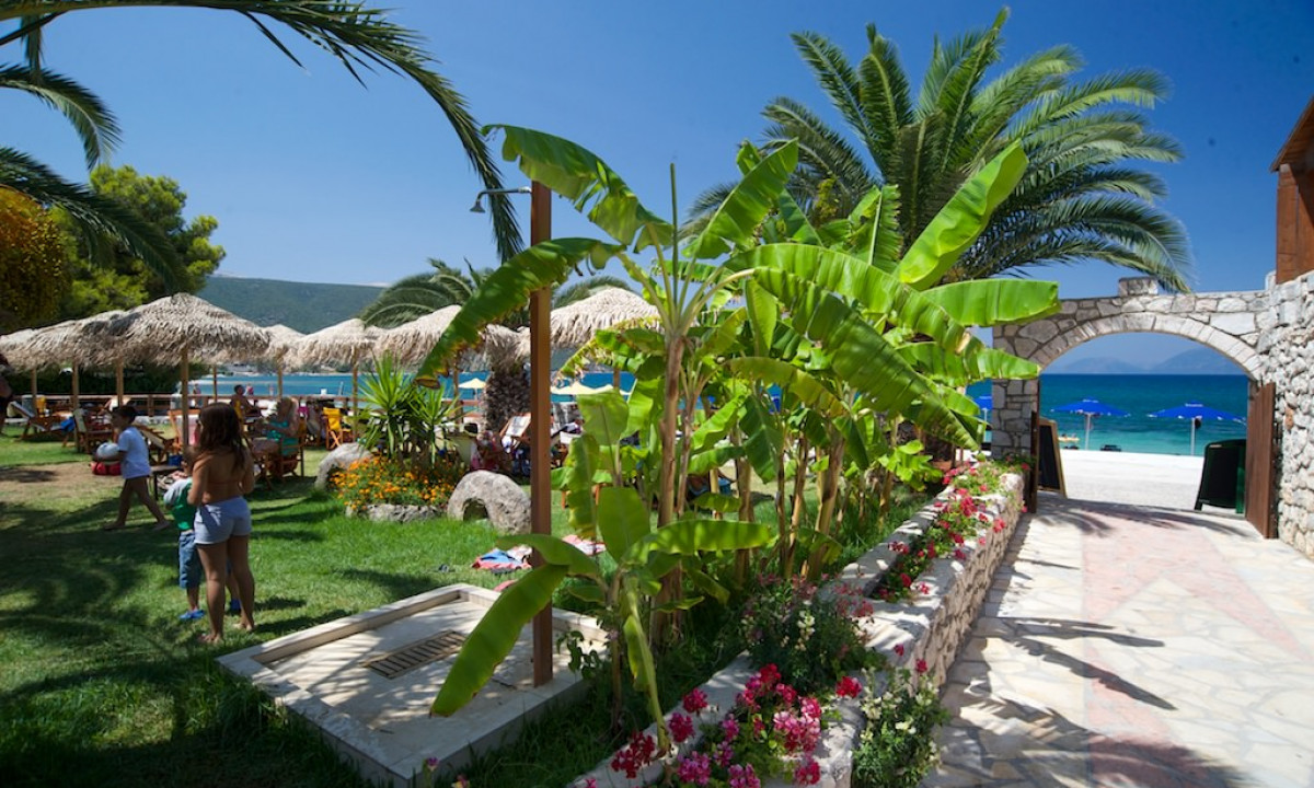 People relaxing and walking near a beach side café with palm trees and umbrellas, overlooking the ocean at Karavomilos Beach