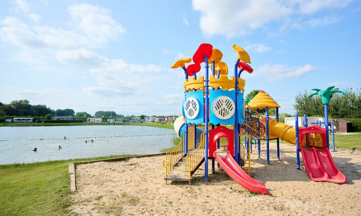 Colorful playground structure with slides and ladders on sandy ground, adjacent to a lake with swimmers at Betuwe