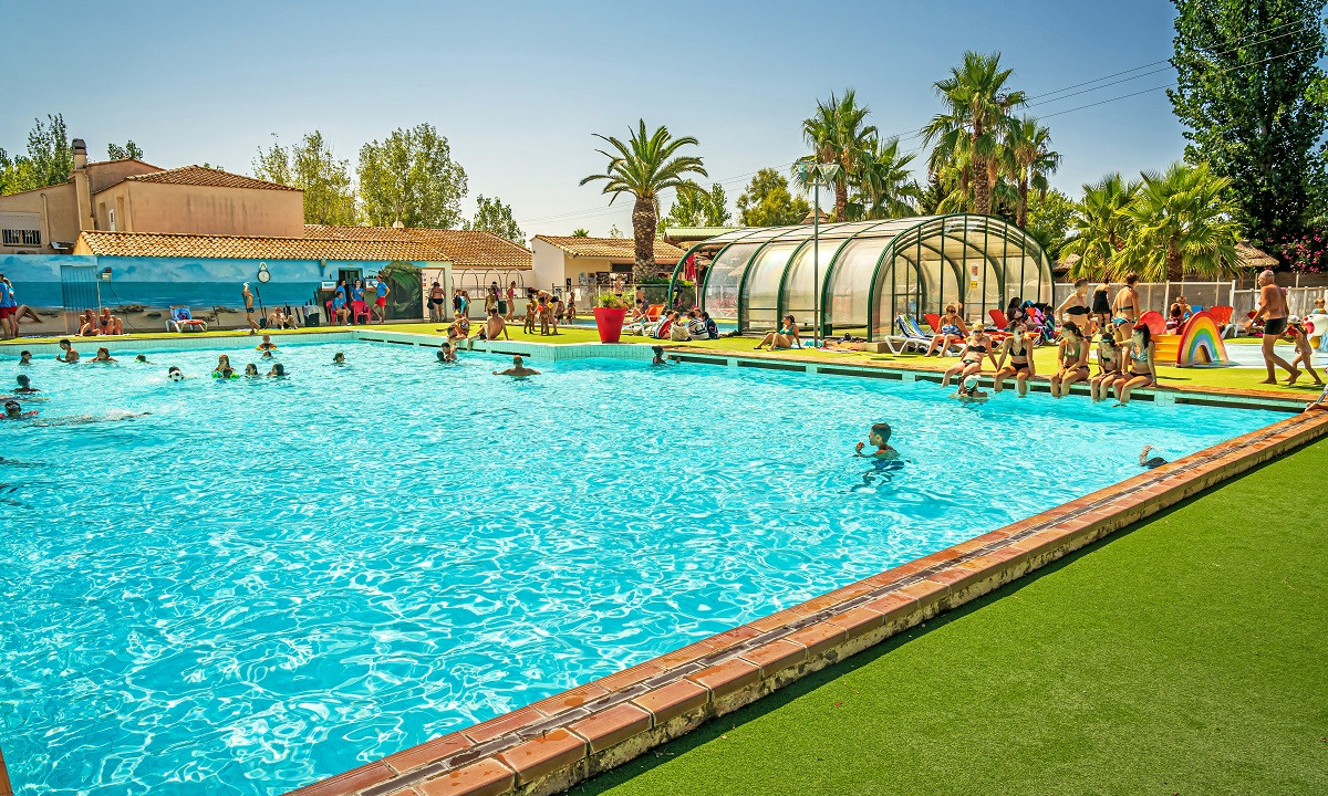 People swimming in a large outdoor pool, while others relax by the poolside; palm trees and buildings surround the area at Le Clos Virgile