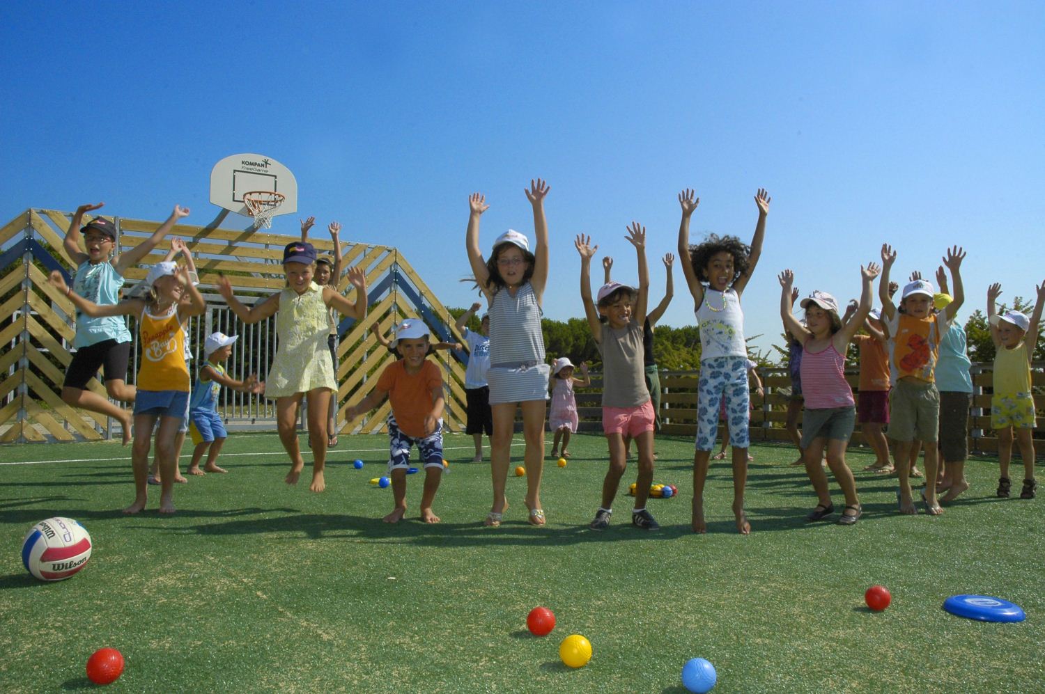 Children jump with raised hands on a playground, surrounded by scattered balls and a basketball hoop at Bon Port