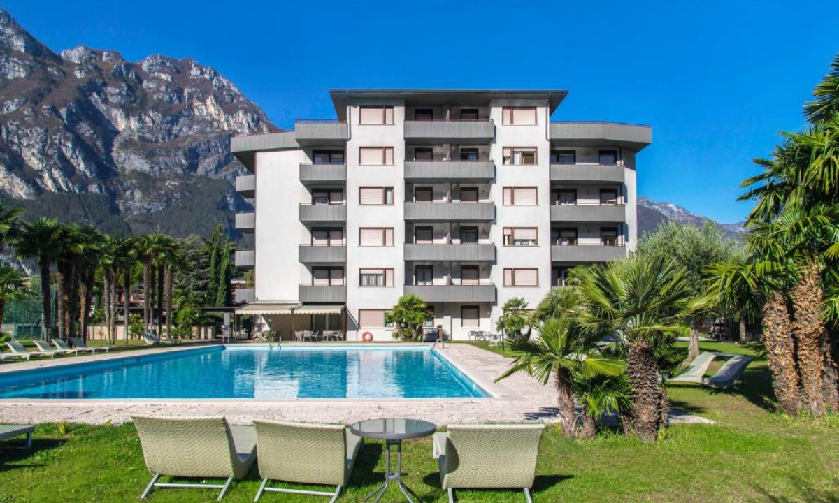 A multi-story building stands behind a large swimming pool surrounded by palm trees and mountains in the background at Residence Monica