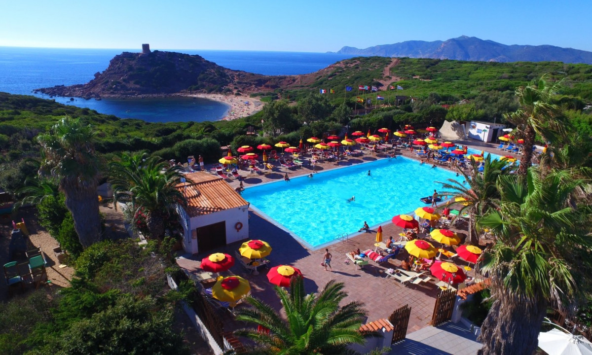 Swimming pool surrounded by people lounging under colorful umbrellas, overlooking a lush, hilly landscape near a coastal bay at Torre del Porticciolo Glamping Village