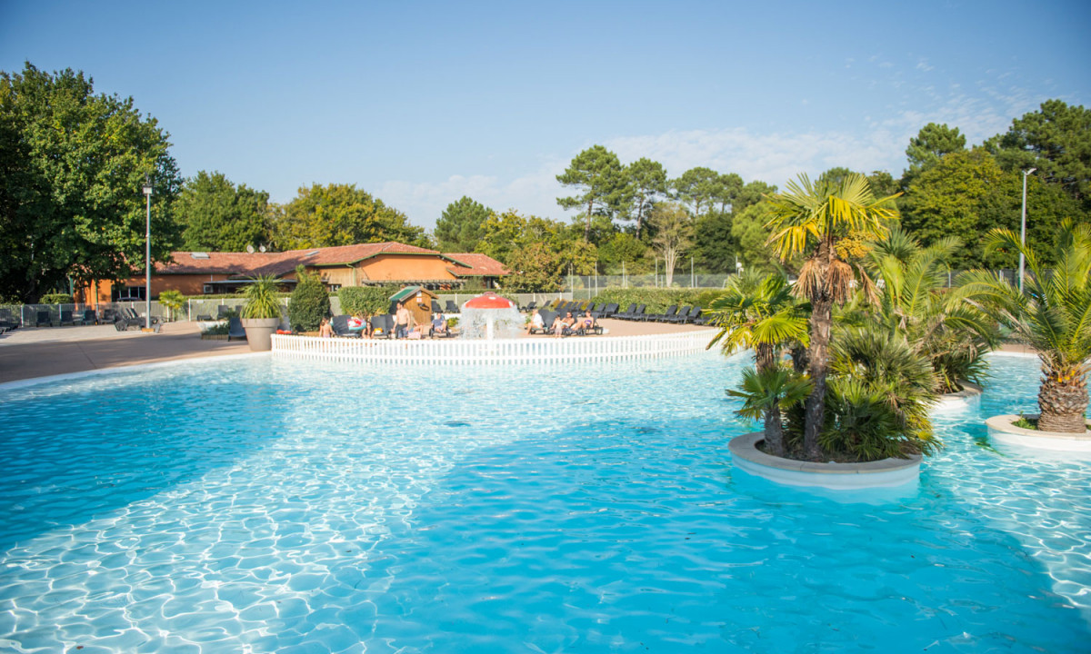Crystal-clear pool, water splashing from mushroom fountain, surrounded by sunbathers, palm trees, and rustic buildings at Domaine de la Forge