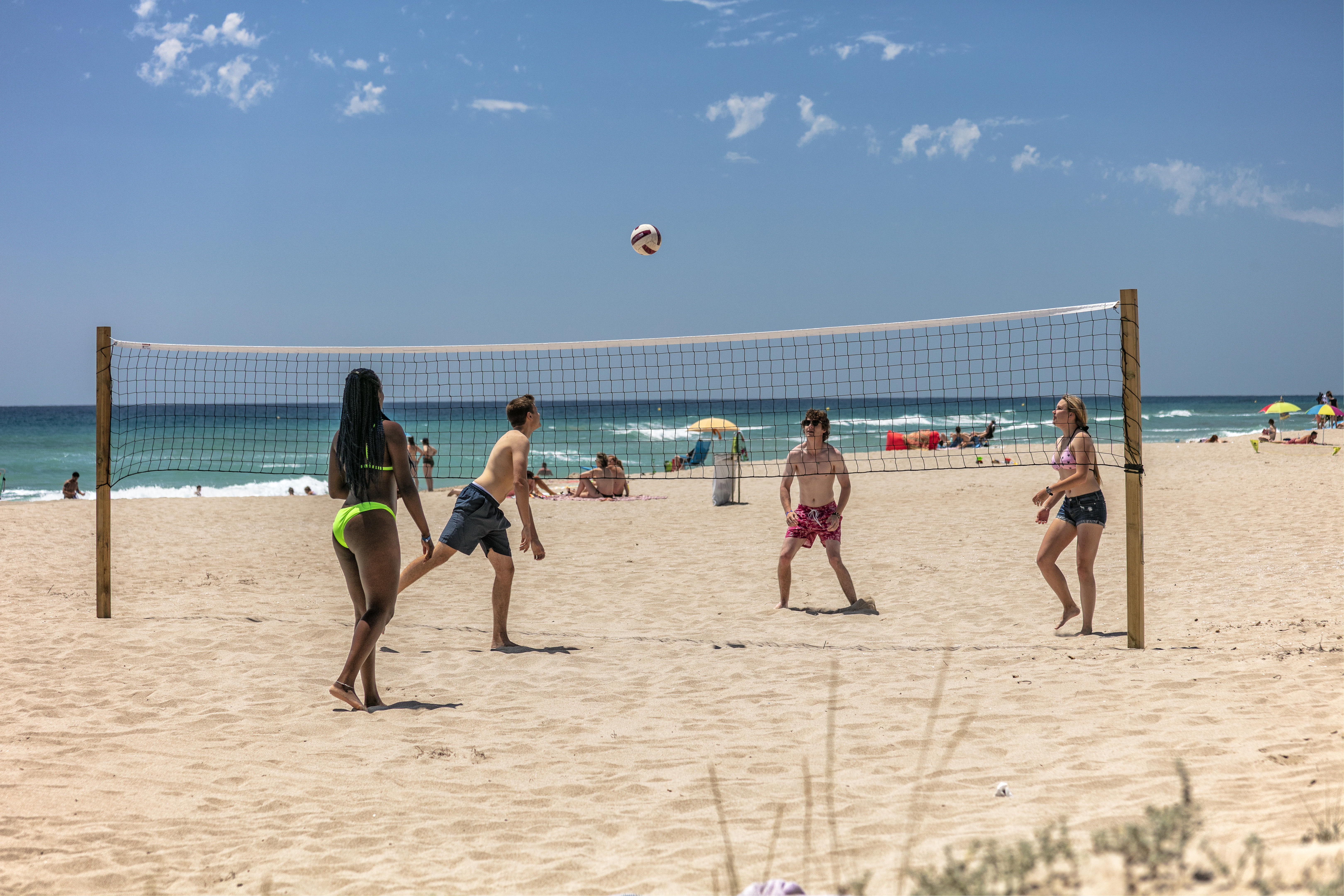 Four people play beach volleyball under a clear sky, with the ocean and sunbathers in the background at Gavina