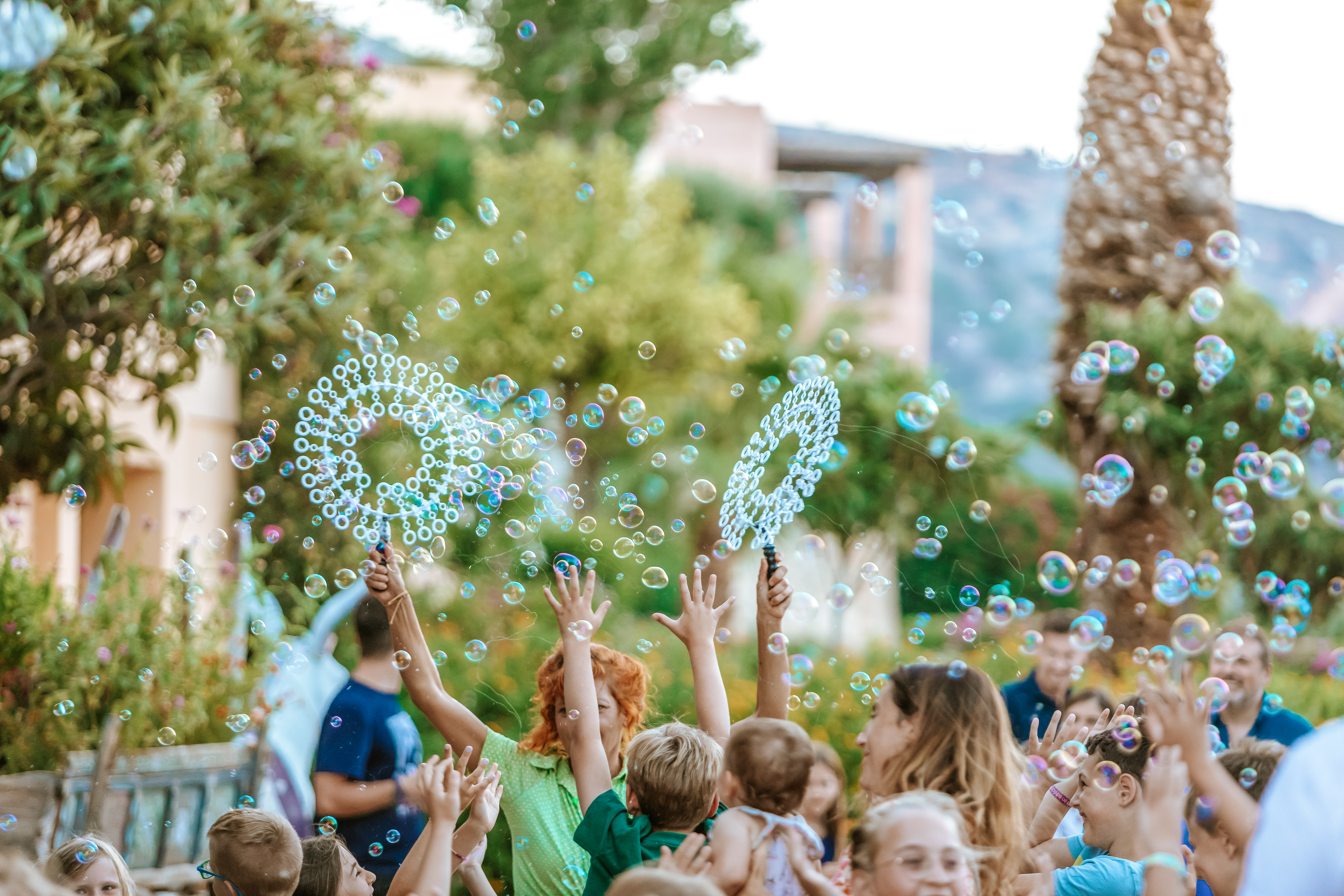 Children joyfully blowing bubbles in the air, surrounded by trees and adults at Candia Park Village
