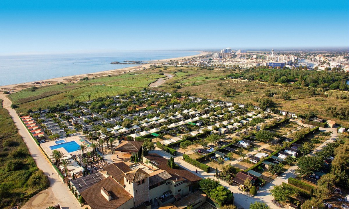 A campsite with organized rows of tents and structures, adjacent to a pool, near a beach and coastal townscape at Castell Mar