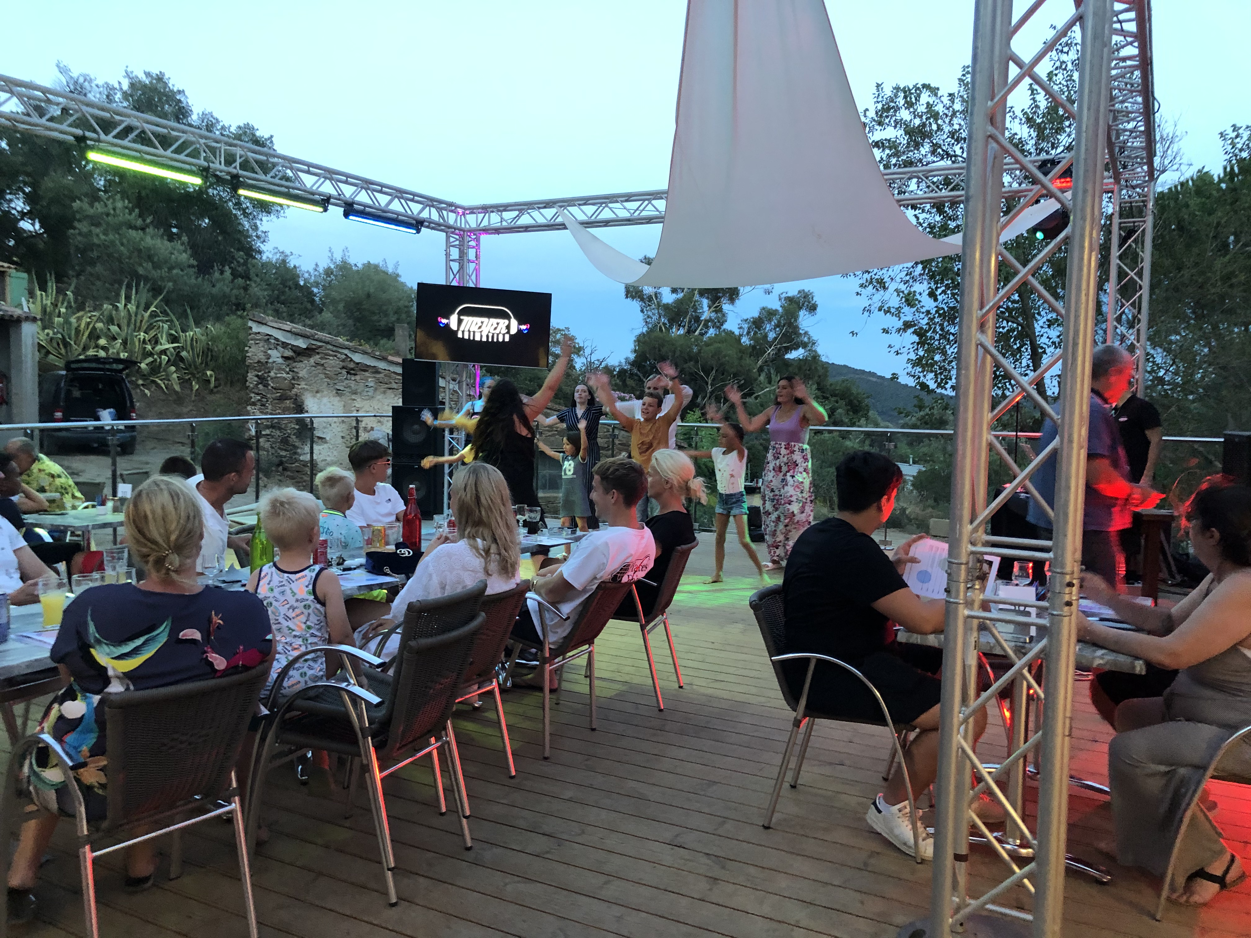 People seated around tables watching children dance on an outdoor stage with a large screen, surrounded by trees at Domaine de Miremer