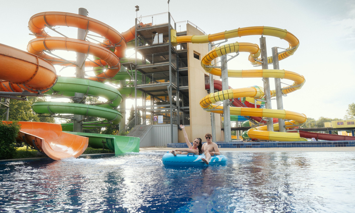 Two people ride a blue inflatable in a pool, surrounded by tall, colorful water slides at Terme Ptuj