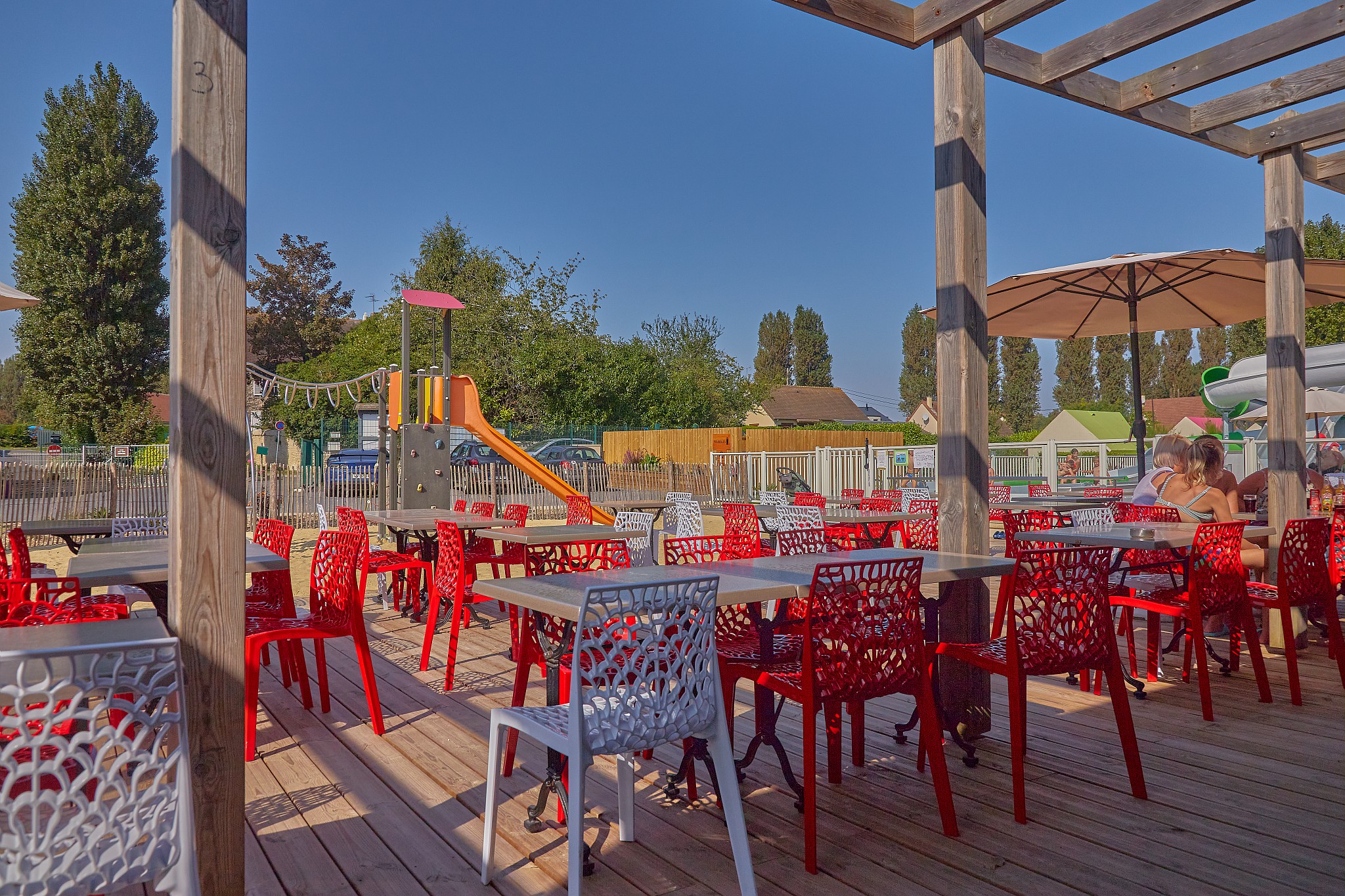 Colorful outdoor seating area with red and white chairs, nearby playground, under wooden pergola at Marvilla Parks Riva Bella
