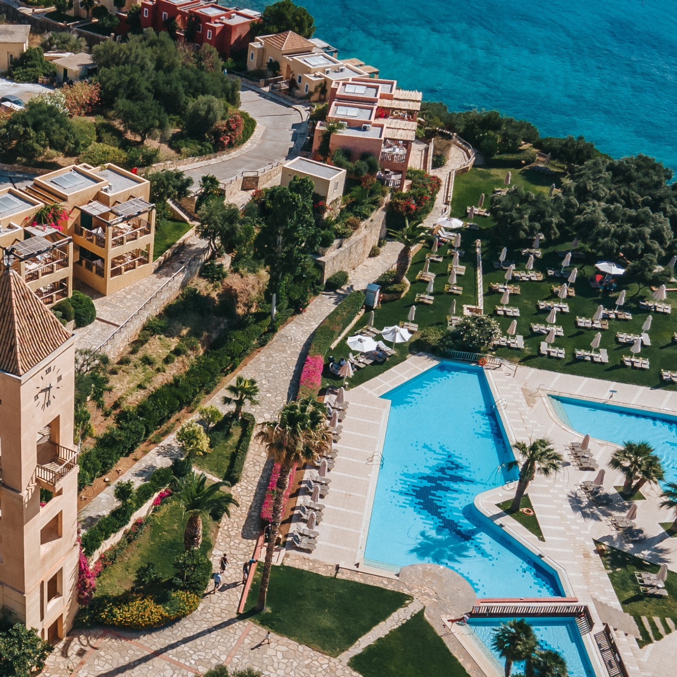 Pool surrounded by lounge chairs and umbrellas, situated near well-manicured gardens and colorful buildings, overlooking the sea at Candia Park Village
