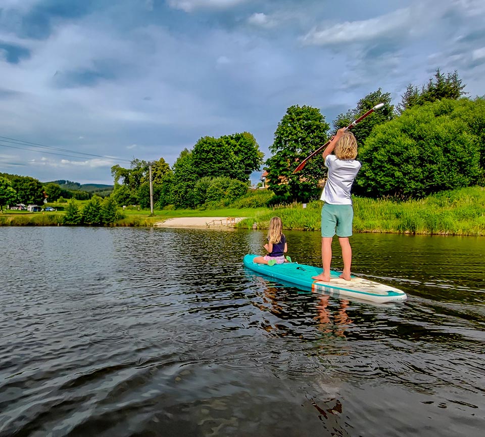 Two children are paddleboarding on a calm lake, surrounded by lush green trees and a sandy shore at Chvalsiny