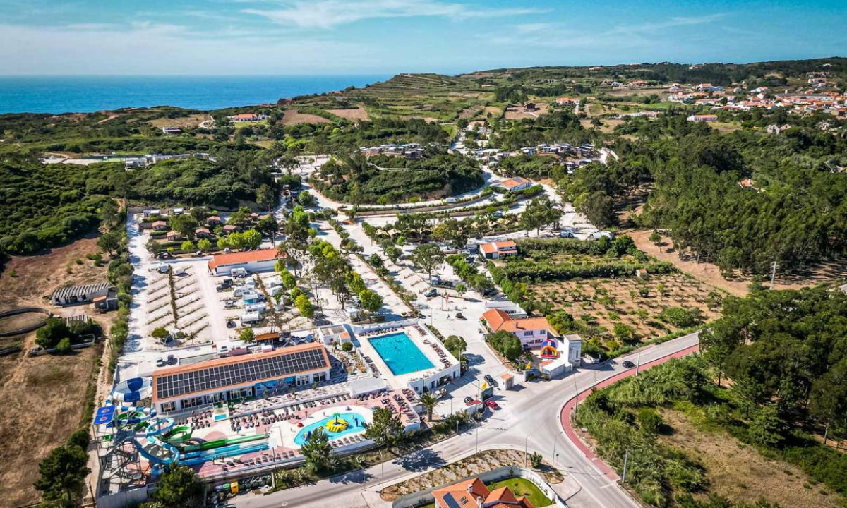 Aerial view of a campsite with multiple pools, water slides, buildings, and parked RVs, surrounded by coastal hills and trees at Yelloh! Colina do Sol
