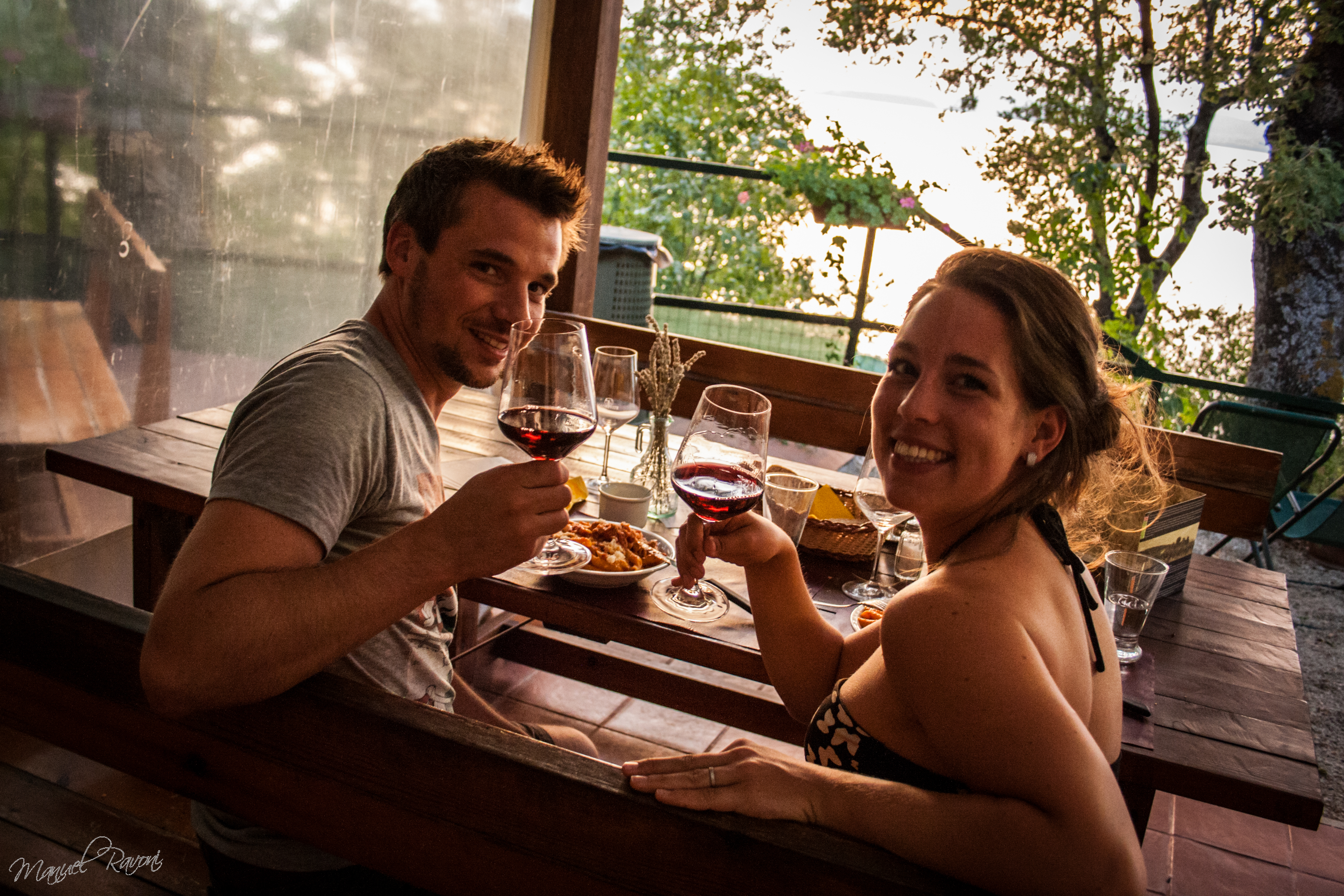 Couple toasting with wine glasses, enjoying a meal together at a wooden picnic table, overlooking scenic greenery at Cerquestra