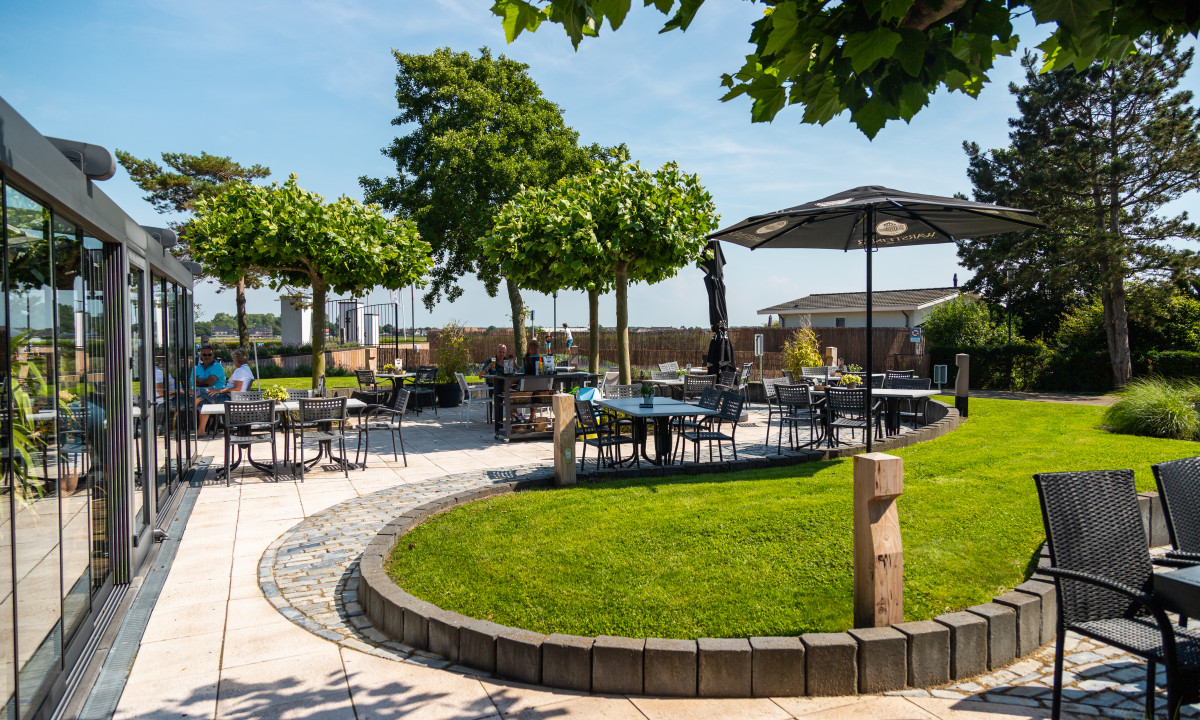Courtyard tables under trees, people dining and conversing, with a lush, green lawn and clear blue sky at Europarcs Parc du Soleil
